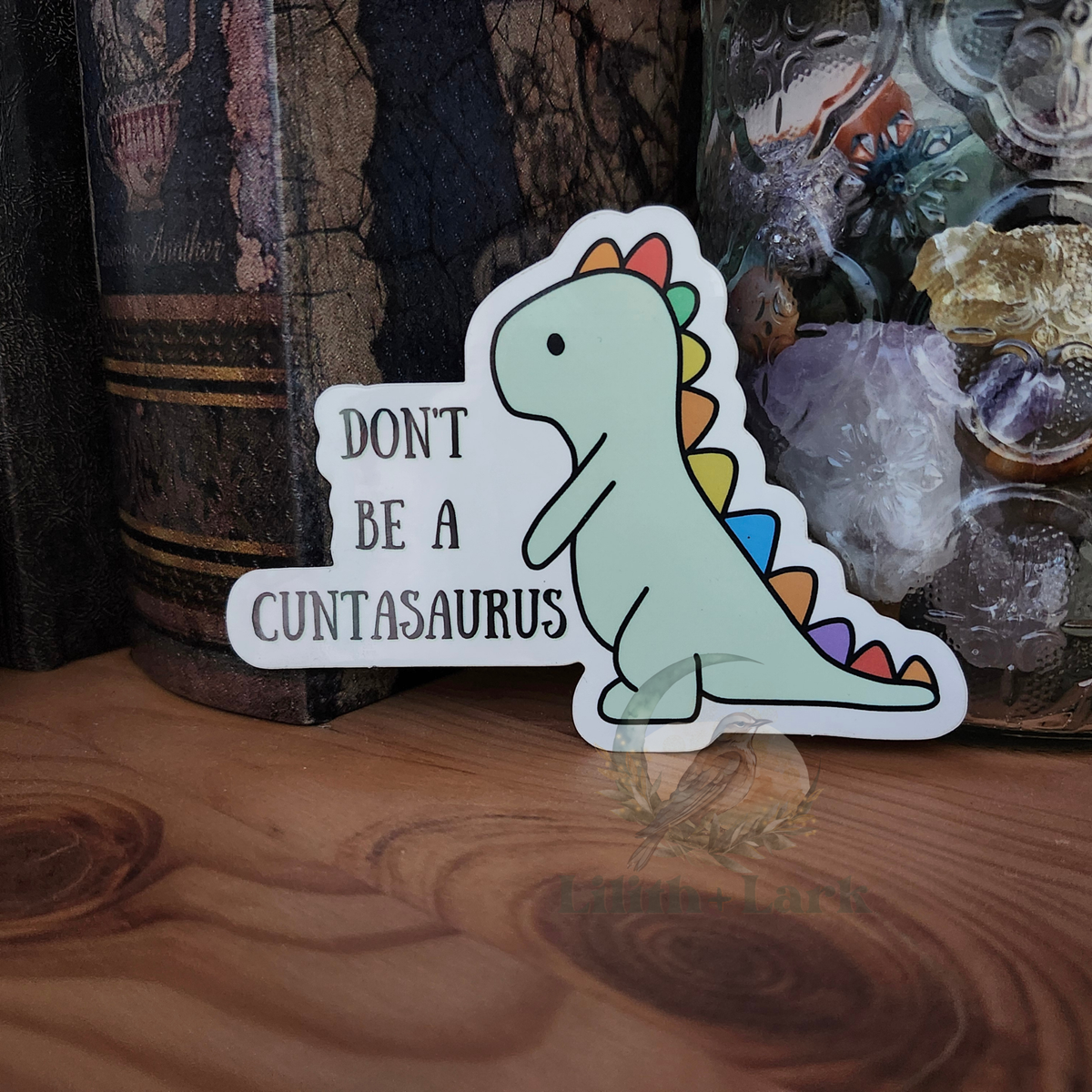 Sticker with a dinosaur design and text that says "Don't be a cuntasaurus" on a wooden surface