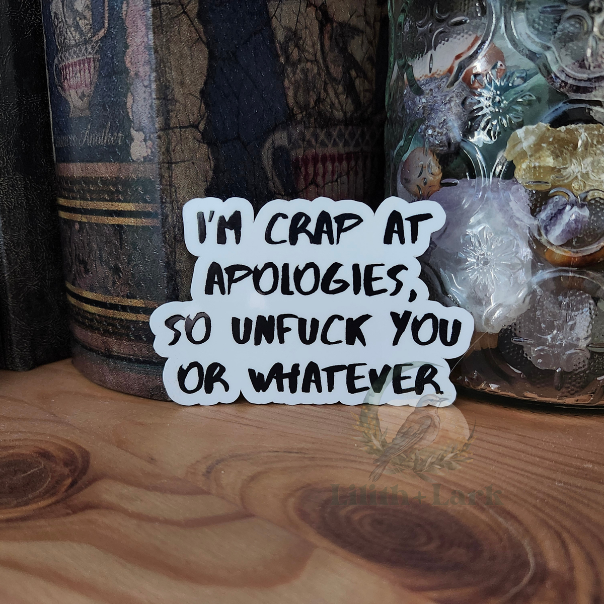 Sticker with the text "I'm crap at apologies, so unfuck you or whatever" on a wooden surface with decorative items in the background